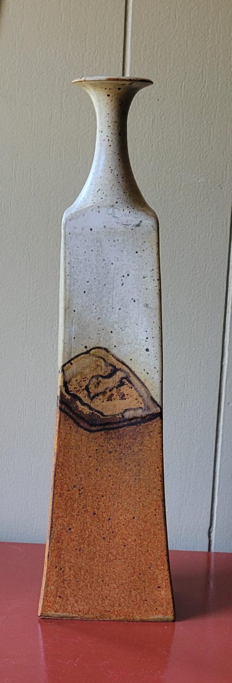 May include: Tall, rectangular ceramic vase with a narrow neck. The upper portion is off-white with black speckles, transitioning to a brown, textured lower section. The vase has a unique, organic shape and is likely handmade.