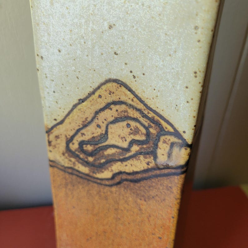 May include: A tall, ceramic vase with a rectangular shape. The top portion is a light beige color, transitioning to a warm orange-brown at the base. A dark brown, stylized mountain design is featured on the upper section.