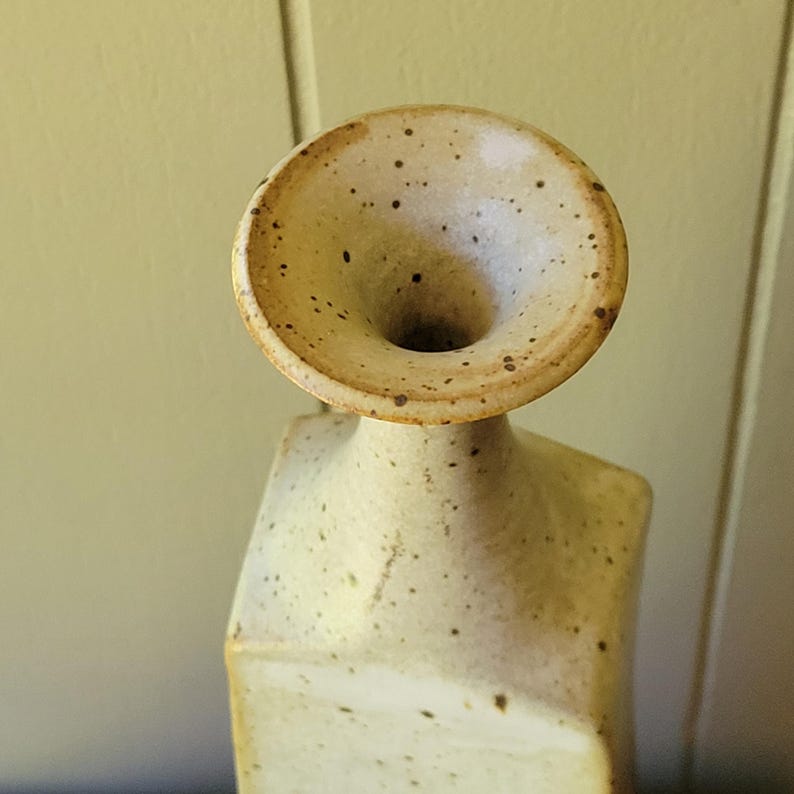 May include: A handcrafted ceramic vase with a unique, abstract design. The vase features a light beige color with dark brown speckles and a flared, funnel-shaped top. The body has a geometric, angular shape.