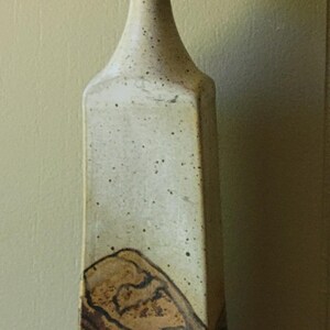 May include: Tall, rectangular ceramic vase with a speckled, light-colored finish and a brown base. The vase holds a sprig of dried, round, beige seed pods. A decorative piece for home decor.