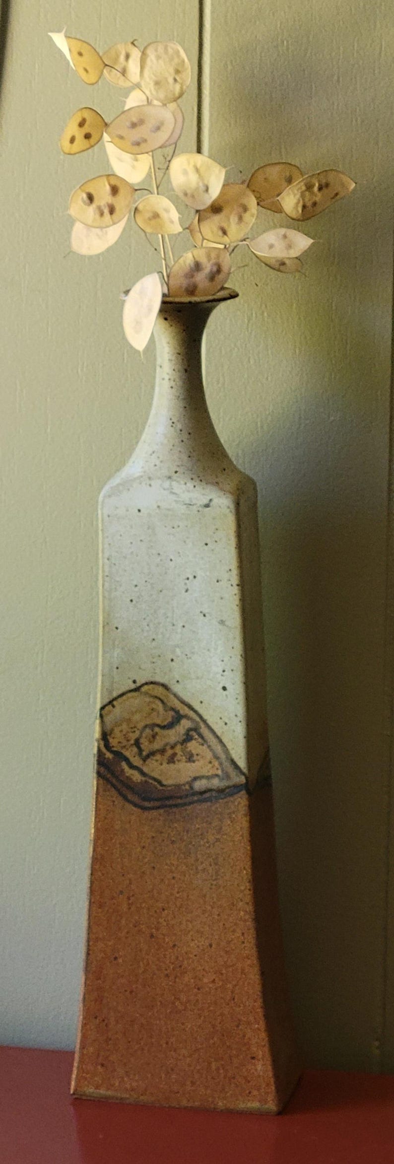 May include: Tall, rectangular ceramic vase with a speckled, light-colored finish and a brown base. The vase holds a sprig of dried, round, beige seed pods. A decorative piece for home decor.