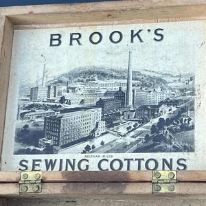 May include: A rectangular wooden box featuring a black and white illustration of a factory complex on the lid. The text BROOK'S is printed above the image, and MELTHAM MILLS SEWING COTTONS is printed below. The box has two brass-colored metal hinges.