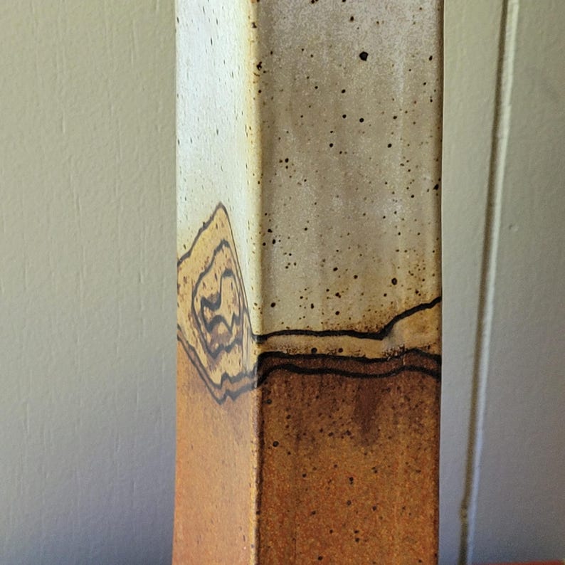 May include: A tall, rectangular ceramic vase with a unique design. The vase features a gradient of colors, transitioning from a warm brown at the base to a speckled beige at the top. The design includes a series of brown, organic shapes and lines.