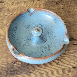 May include: A round ceramic ashtray featuring a speckled light blue glaze with a brown rim. The center of the ashtray has a small, raised circular post, and the outer edge includes three distinct notches for resting items, showcasing a rustic finish.