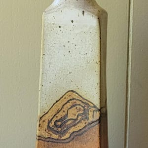 May include: Tall, rectangular ceramic vase with a narrow neck. The upper portion is off-white with dark speckles, transitioning to a brown base with a textured, layered design. A unique piece for home decor.