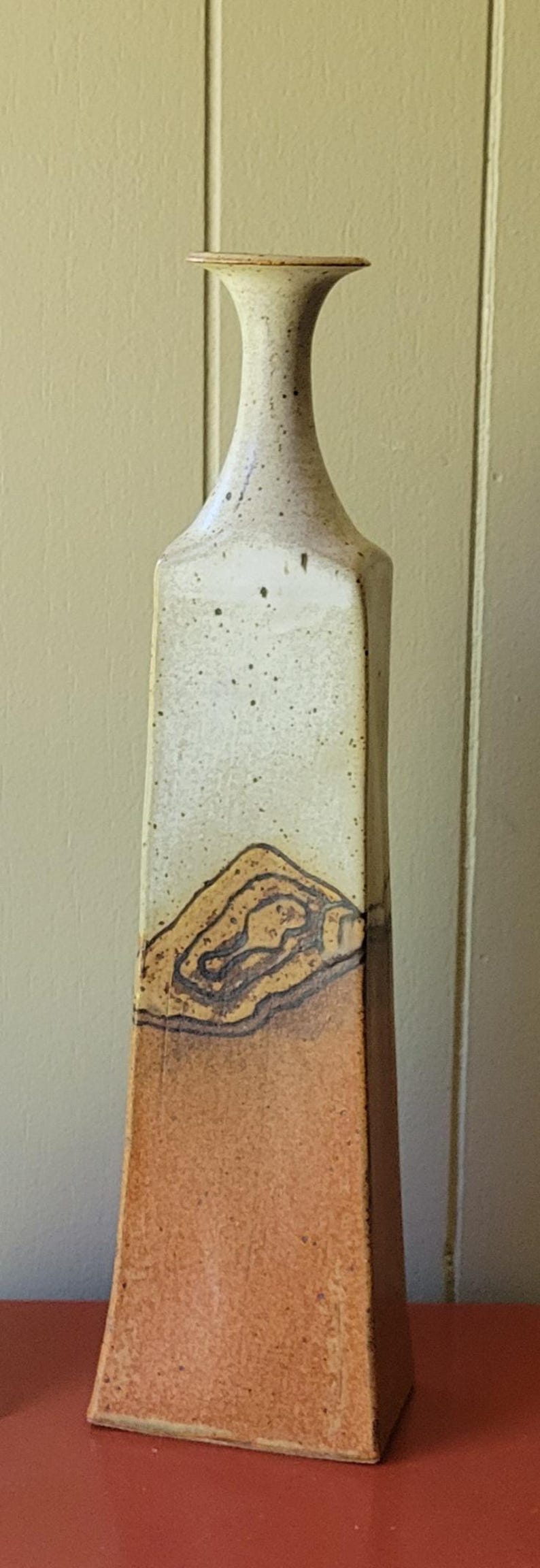 May include: Tall, rectangular ceramic vase with a narrow neck. The upper portion is off-white with dark speckles, transitioning to a brown base with a textured, layered design. A unique piece for home decor.