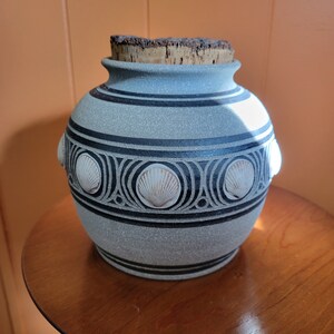 May include: A gray ceramic jar with a cork lid and a decorative design of seashells. The jar has a textured surface and a black and white design.