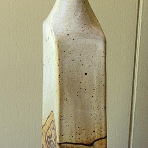 May include: Tall, ceramic vase with a square base and a flared top. The vase has a speckled, off-white finish with brown accents and a band of decorative swirls. The vase stands approximately 12 inches tall.