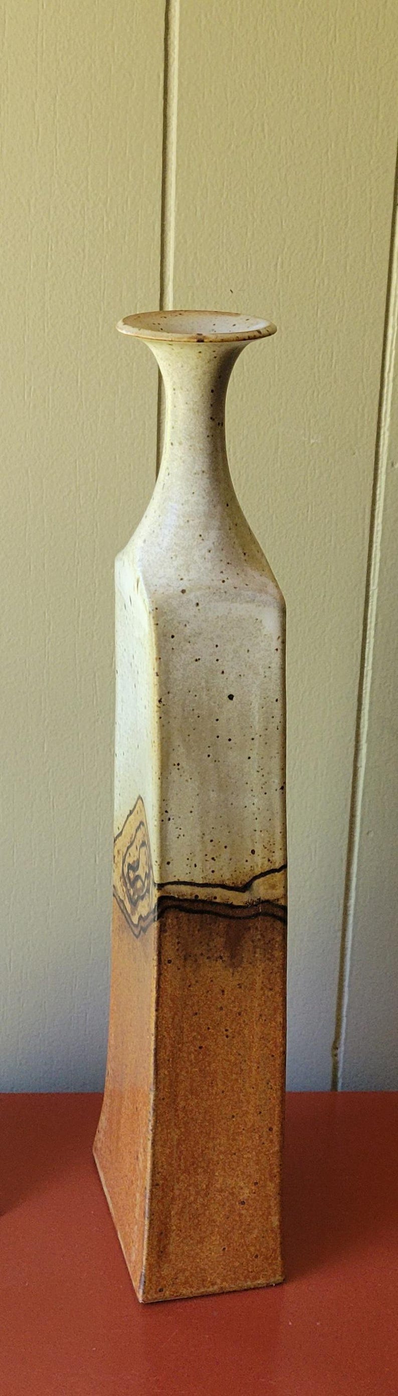 May include: Tall, ceramic vase with a square base and a flared top. The vase has a speckled, off-white finish with brown accents and a band of decorative swirls. The vase stands approximately 12 inches tall.