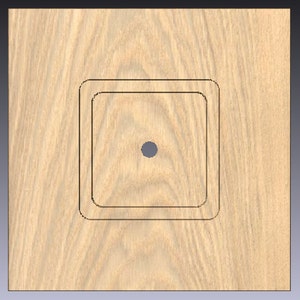May include: A light-colored wooden square with a centered, circular hole. Two black square outlines are etched into the wood, creating a recessed design. The wood grain is visible.