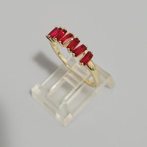 May include: A gold-colored ring featuring a series of rectangular, vibrant red gemstones. The ring is displayed on a clear, angled stand, showcasing its design and color contrast. The gemstones are evenly spaced along the ring's band.