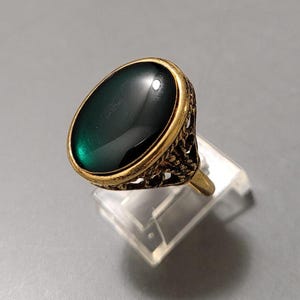 May include: A gold-toned ring featuring a large, oval-shaped dark green cabochon stone set in a bezel. The band and setting exhibit an intricate, openwork filigree metal design that wraps around the sides of the central gemstone.