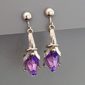 May include: A pair of silver-toned drop earrings. Each earring features a faceted, teardrop-shaped purple gemstone suspended from a decorative silver element. The earrings have a post-and-ball closure.