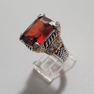 May include: A silver ring with a large, rectangular, red gemstone. The ring has gold accents and small clear stones around the main stone. The band has a textured design.