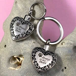 May include: Two silver heart-shaped cremation keychains with a paw print and the inscription "Always in my Heart".