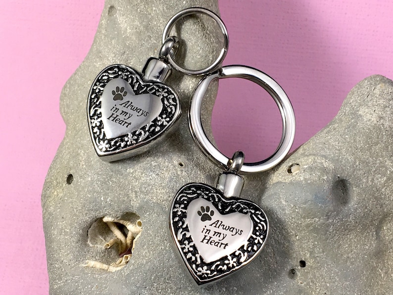 May include: Two silver heart-shaped cremation keychains with a paw print and the inscription "Always in my Heart".