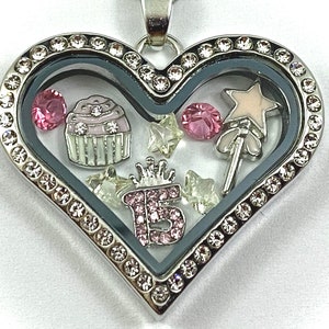 May include: Silver heart-shaped locket with a clear glass insert. The locket is decorated with rhinestones and contains charms including a cupcake, a star, a wand, and a pink number 15.