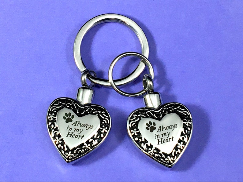 May include: Silver keychain with two heart-shaped cremation urns. Each urn has a paw print and the inscription "Always in my Heart".