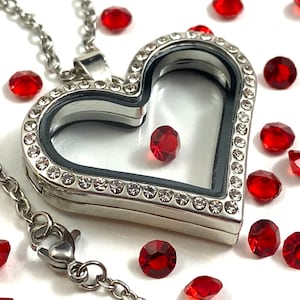 May include: A silver heart-shaped locket necklace with a clear glass insert and a small red gem inside. The locket is surrounded by a row of clear rhinestones. The necklace is on a silver chain with a lobster clasp.