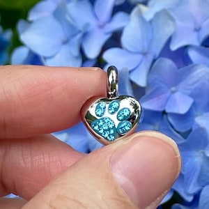 May include: A heart-shaped pendant with a paw print design. The pendant is silver-colored with a blue paw print. The background is a cluster of blue hydrangea flowers.