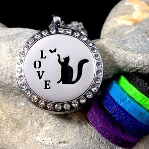 May include: A silver-toned pendant with a circular face, adorned with clear crystals around the edge. The face features the word "LOVE" and a black cat silhouette reaching for a small bird. A stack of colorful felt pads is visible.