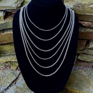 May include: A collection of silver-colored chain necklaces of varying lengths displayed on a black velvet bust. The necklaces are delicate and hang in a layered arrangement, showcasing different chain styles. The background features a stone wall.