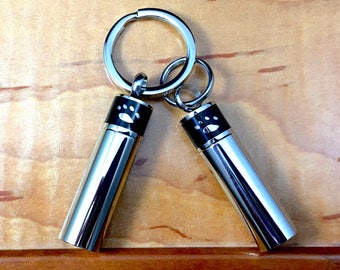 May include: Two silver keychains with black paw print accents. Each keychain has a silver ring and a cylindrical container with a screw-on lid.