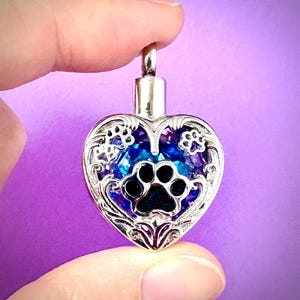 May include: Silver heart-shaped pendant with a paw print design. The pendant features a blue and purple crystal-like center and a decorative border. A small silver loop is at the top for a chain.