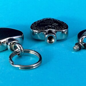 May include: Three silver heart-shaped cremation keychains with a screw-on lid. One keychain has the inscription "In My Heart Always" with a paw print.