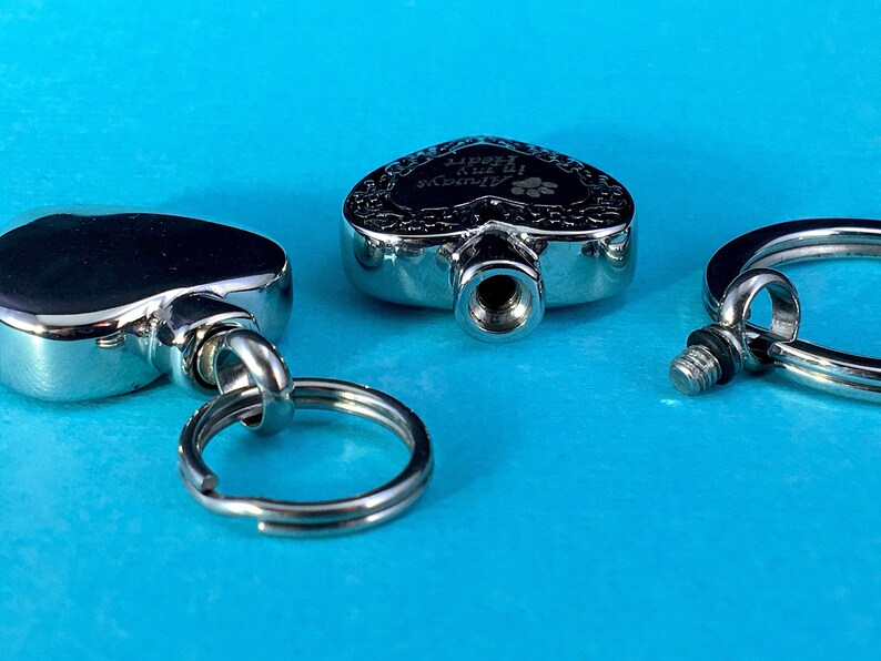 May include: Three silver heart-shaped cremation keychains with a screw-on lid. One keychain has the inscription "In My Heart Always" with a paw print.
