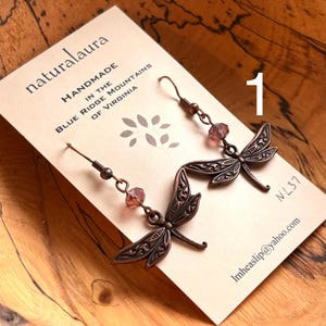 May include: Copper-colored dragonfly earrings with pink beads. The earrings are handmade in the Blue Ridge Mountains of Virginia. The earrings are on a cream-colored card with the text "naturalaura".