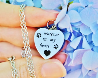 May include: Silver heart-shaped pendant necklace with a chain. The pendant has the inscription "Forever in my heart" and two paw prints on either side.