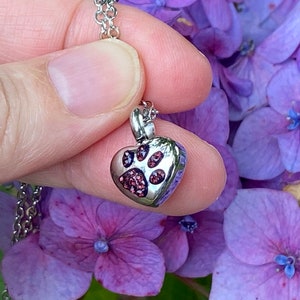 May include: A silver heart-shaped pendant necklace with a paw print design. The pendant is filled with small pink crystals. The necklace is on a silver chain.
