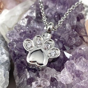 May include: Silver paw print cremation necklace with clear cubic zirconia stones. The necklace is on a bed of purple amethyst crystals.