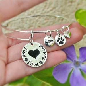 May include: Silver bangle bracelet with charms. The charms are a heart, a paw print, and a round charm with the words "Live, Love, Rescue".