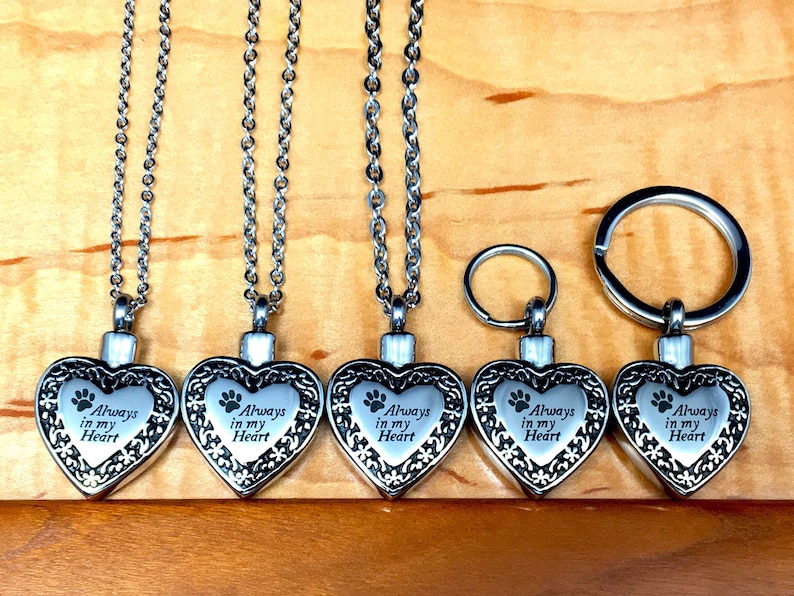 May include: Five silver heart-shaped cremation pendants with a paw print and the inscription "Always in my Heart". One pendant is attached to a silver key chain.
