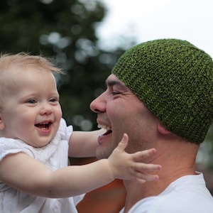 Puede incluir: Un hombre con un gorro de punto verde sonríe a una niña pequeña que lleva un vestido blanco. La niña está sujetando la mano del hombre y lo está mirando.
