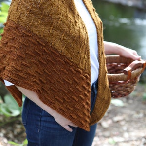 May include: A brown knitted shawl with a textured pattern. The shawl is draped over a person's shoulders and is worn with a white shirt and blue jeans.