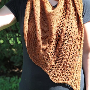 May include: A brown knitted shawl with a mix of solid and openwork patterns. The shawl's colour is a warm, earthy tone. The design includes a triangular shape, draped over a dark top. The openwork sections add a delicate texture to the overall design, creating a stylish accessory.