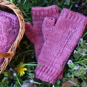 May include: A pair of pink knitted fingerless gloves with a cable knit pattern. The gloves are lying on a bed of green grass and small purple flowers.