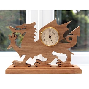 May include: Wooden desk clock in the shape of a dragon. The clock has a gold-colored face with black numbers and hands. The dragon is carved from light brown wood and stands on a rectangular base. The clock is set against a window.
