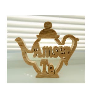 May include: Wooden decorative sign shaped like a teapot. The light brown sign has the words "Amser Te" cut out in the middle. The sign is positioned in front of a window with horizontal blinds.