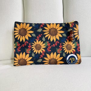 May include: A rectangular, navy blue zippered pouch with a sunflower pattern. The sunflowers are yellow and brown, with red and green leaves. A small gold zipper pull has brown beads. The pouch has a small circular logo that says "ATB".