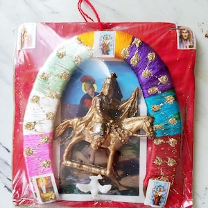 May include: A religious icon featuring a gold-colored statue of a person on horseback, set against a backdrop of a framed image and a rainbow-colored horseshoe. The horseshoe is wrapped in colorful yarn and adorned with gold glitter.