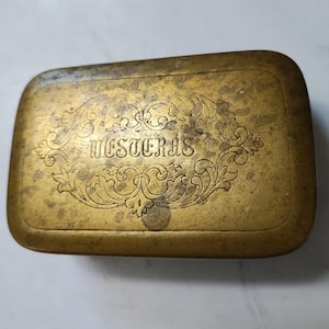 May include: A rectangular, antique brass box with rounded corners. The box features an ornate, engraved floral design with the word "WESTERAS" in the center. The brass has a worn, aged appearance.