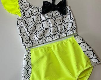 May include: A two-piece outfit for girls, featuring a neon yellow and white crop top with a black bow and a neon yellow high-waisted bottom with a white smiley face pattern.