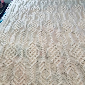 May include: White knitted throw blanket with a cable knit pattern. The blanket has a diamond-shaped design with a repeating pattern of knots.