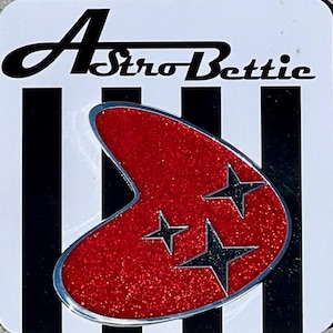 May include: Astro Bettie coaster with a red, glittery boomerang design and three black star accents. The coaster has a silver trim and is set against a black and white striped background.