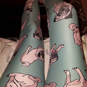 May include: A pair of teal blue leggings with a repeated pattern of pugs in various poses. The pugs are a light pink color with black noses and eyes.