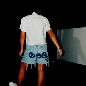 May include: A person wearing a white button-down shirt and denim shorts with blue smiley face patches. The person is wearing white sneakers.